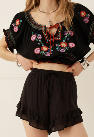A woman wearing a black embroidered crop top with colorful floral patterns and matching black ruffled shorts, standing against a light beige wall.